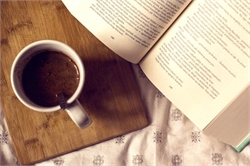 [ai] A white coffee cup filled with dark coffee sits on a wooden cutting board next to an open book with printed text. The background features a light-colored patterned fabric.